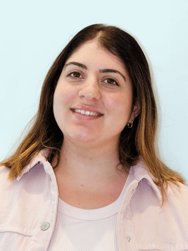 A person with shoulder-length brown hair smiles slightly. They are wearing a light pink shirt over a white top, with small hoop earrings. The background is a plain light blue.